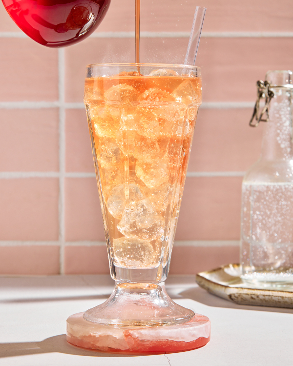 A parfait glass of old-fashioned cream soda on ice shown with a bottle of soda and pouring strawberry simple syrup from a pitcher into the glass