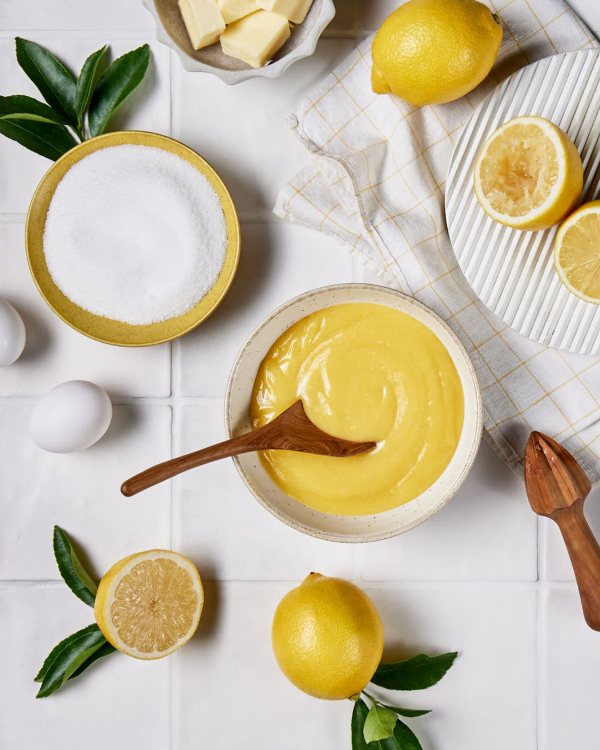 Lemon Curd A bowl of lemon curd shown on a tiled counter with whole and halved lemons, a lemon juicer, and a bowl of granulated sugar.
