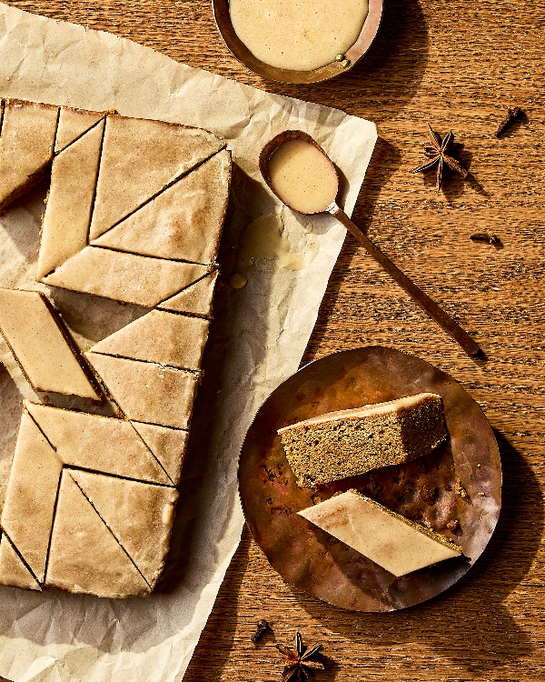 Rows of spiced cake bars cut into diamond shapes, topped with a smooth glaze. One bar is broken in half, revealing a moist and crumbly interior. The warm brown background looks rustic and cozy