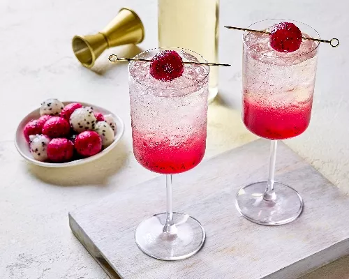 Two glasses of vibrant dragon fruit spritzer served over ice, with a pink gradient from the fruit at the bottom. Each glass is garnished with a skewered dragon fruit ball. A plate of red and white dragon fruit balls, a gold jigger, and a bottle are visible in the background.