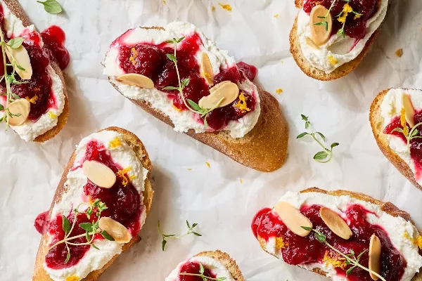 A close-up view of several crostini topped with whipped feta cheese, cranberry chutney, slivered almonds, and fresh thyme.